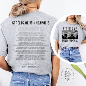 May include: A light gray t-shirt featuring the text "STREETS OF MINNEAPOLIS" and a poem by Bruce Springsteen. The shirt also has a graphic of a protest. The brand logo is visible on the bottom right.