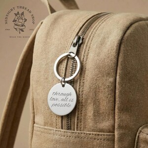 May include: A silver-colored round keychain with the inscription "through love, all is possible" hangs from a backpack zipper. The backpack is a light brown color, and the keychain is attached to a silver ring.
