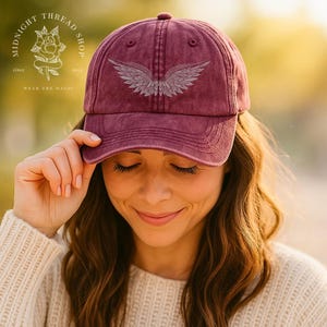 May include: A maroon baseball cap with a distressed finish, featuring a detailed silver wing design. The cap has a curved brim and a top button. The Midnight Thread Shop logo is visible in the background.