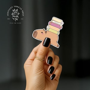 May include: A die-cut vinyl sticker featuring a tan capybara with a stack of three pastel-coloured books balanced on its back. The books are light yellow, pink, and light green, each accented with a small white flower detail on the spine.