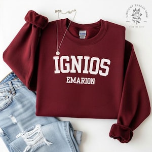 Puede incluir: Una sudadera granate con la palabra "IGNIOS" encima de "EMARION" en letras blancas. La sudadera tiene cuello redondo y puños. Un collar plateado con un pequeño colgante está encima de la sudadera, que está sobre unos vaqueros azules.