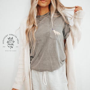 May include: A gray t-shirt with a pocket featuring a white fox design. The shirt is part of a casual outfit, paired with light gray sweatpants and a cream-colored cardigan. The text "MIDNIGHT THREAD SHOP" and a rose logo are visible.