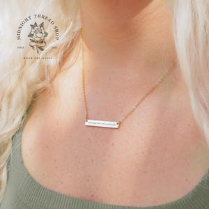 May include: A gold-colored necklace with a rectangular pendant. The pendant has the inscription "through love, all is possible". The necklace is worn on a person with blonde hair and a green top. The shop logo is visible in the upper left corner.