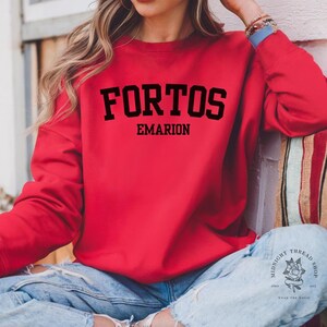 Puede incluir: Una sudadera roja con la palabra "FORTOS" en letras negras en negrita sobre "EMARION". La sudadera es de color rojo liso y tiene cuello redondo. Las mangas son largas y el material parece ser una tela suave.