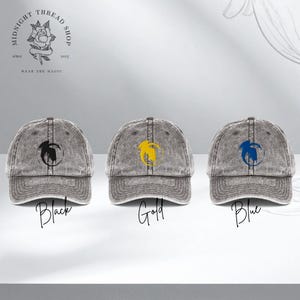 Puede incluir: Tres gorras de béisbol grises con un acabado desgastado. Cada gorra presenta un diseño circular con un gráfico de pájaro en negro, dorado y azul. Las palabras "Black", "Gold" y "Blue" están escritas debajo de cada gorra.