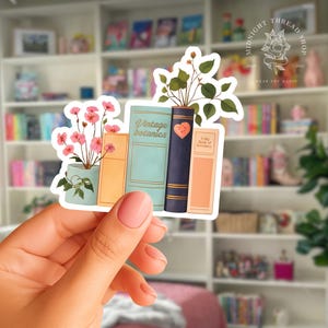 May include: A die-cut sticker featuring a collection of books and potted plants. The books include a light blue volume labeled Vintage botanics, a dark blue book with a heart, and a peach book labeled A Big Book of greenery. Pink flowers and green leaves emerge from the pots.