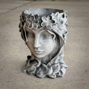 May include: A gray concrete planter shaped like a woman's face, adorned with a floral crown. The detailed sculpture features closed eyes and flowing hair, with a textured finish. This decorative item is suitable for indoor or outdoor use.