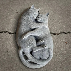 May include: A gray cat sculpture depicting two cats embracing. The cats are in a close embrace, with their eyes closed. The sculpture is made of a stone-like material and has a textured surface. The cats' tails are intertwined.