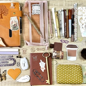 May include: A collection of stationery items including two notebooks, a transparent pencil case, various pens, markers, stickers, paper clips, a key-shaped bookmark, a small patterned pouch, a reading list, and a brown scrunchie arranged on a surface.