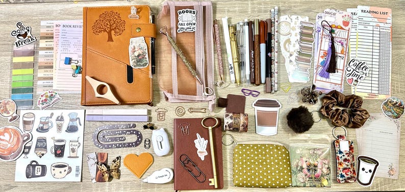 May include: A collection of stationery items including two notebooks, a transparent pencil case, various pens, markers, stickers, paper clips, a key-shaped bookmark, a small patterned pouch, a reading list, and a brown scrunchie arranged on a surface.