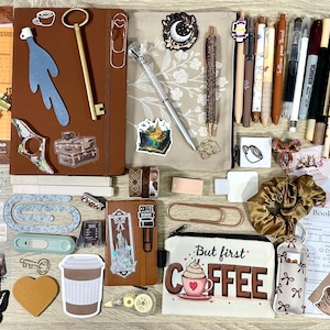 May include: A collection of stationery items including a brown notebook, a pencil case with the text But first COFFEE, various pens, stickers, paper clips, a gold key, a scrunchie, sticky notes, and book review cards arranged on a flat surface.