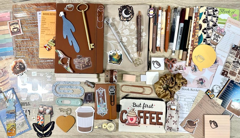 May include: A collection of stationery items including a brown notebook, a pencil case with the text But first COFFEE, various pens, stickers, paper clips, a gold key, a scrunchie, sticky notes, and book review cards arranged on a flat surface.