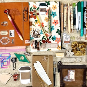 May include: A collection of stationery items including two notebooks, various pens, bookmarks, stickers, paper clips, a small brown pouch, a fuzzy heart keychain, and a scrunchie. The items feature floral patterns, coffee motifs, and text like Dream Big.