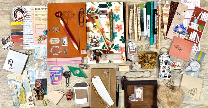 May include: A collection of stationery items including two notebooks, various pens, bookmarks, stickers, paper clips, a small brown pouch, a fuzzy heart keychain, and a scrunchie. The items feature floral patterns, coffee motifs, and text like Dream Big.
