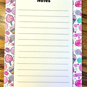 Può includere: Un blocco note rettangolare con un centro bianco che presenta la parola Notes in testo nero sopra linee orizzontali. Il bordo è decorato con un motivo colorato di zucchero filato rosa, fette di torta arcobaleno, lecca-lecca e labbra rosa.
