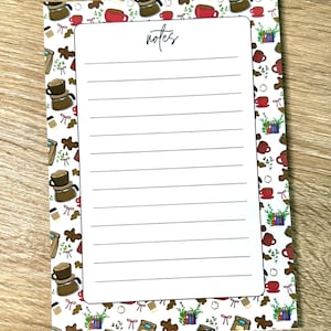 May include: A rectangular notepad with a white center featuring horizontal lines and the word notes at the top. The border is decorated with a repeating pattern of coffee carafes, red mugs, stacks of books, gingerbread cookies, and small bows.