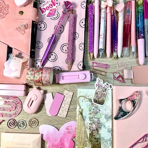 Pu&ograve; includere: Una collezione di articoli di cancelleria rosa che include un'agenda, un astuccio con faccine sorridenti, varie penne con glitter e cristalli, nastro adesivo decorativo floreale, un taglierino rosa, foglietti adesivi a farfalla e graffette.