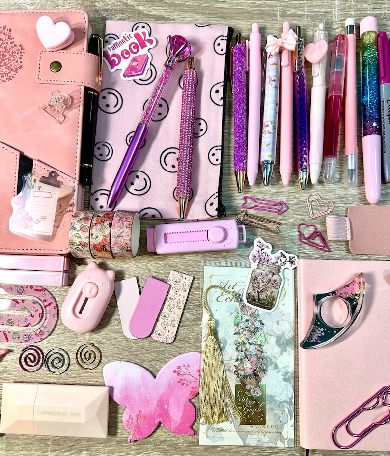 Pu&ograve; includere: Una collezione di articoli di cancelleria rosa che include un'agenda, un astuccio con faccine sorridenti, varie penne con glitter e cristalli, nastro adesivo decorativo floreale, un taglierino rosa, foglietti adesivi a farfalla e graffette.