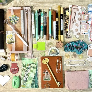May include: A collection of stationery and reading accessories including two journals, various pens, markers, stickers, bookmarks, paper clips, a small pink pouch, a scrunchie, a key-shaped pen, and reading list inserts arranged on a flat surface.