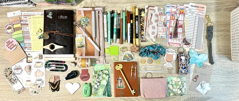 May include: A collection of stationery and reading accessories including two journals, various pens, markers, stickers, bookmarks, paper clips, a small pink pouch, a scrunchie, a key-shaped pen, and reading list inserts arranged on a flat surface.