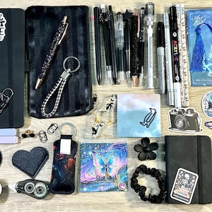 May include: A collection of stationery items including two black notebooks, a black pencil case, various pens, markers, stickers, bookmarks, paper clips, washi tape, a beaded keychain, a scrunchie, and sticky notes arranged on a flat surface.