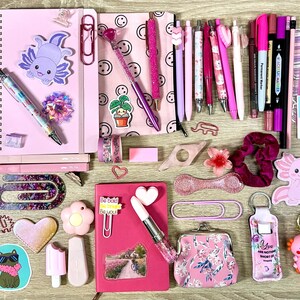 Pu&ograve; includere: Una collezione di articoli di cancelleria che include quaderni a spirale rosa, varie penne, pennarelli e adesivi con motivi di axolotl, gatti e farfalle. Sono inclusi anche graffette, un piccolo portamonete floreale e un portachiavi.