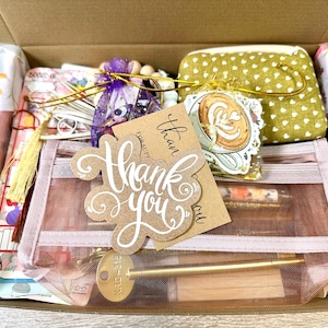 May include: A cardboard gift box containing a pink mesh pencil case with a thank you tag, a gold-coloured metal pen, a small olive green pouch with white hearts, a purple organza bag with wooden beads, and a sticker featuring a coffee cup.