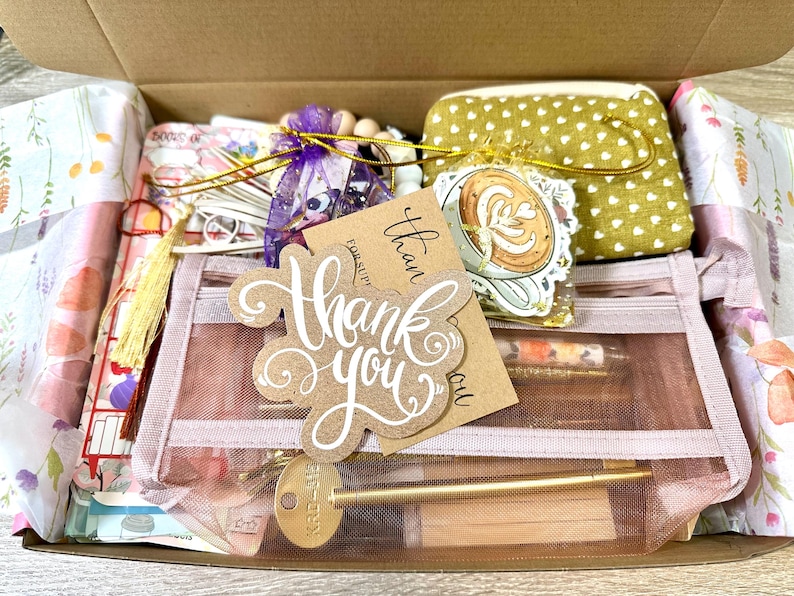 May include: A cardboard gift box containing a pink mesh pencil case with a thank you tag, a gold-coloured metal pen, a small olive green pouch with white hearts, a purple organza bag with wooden beads, and a sticker featuring a coffee cup.