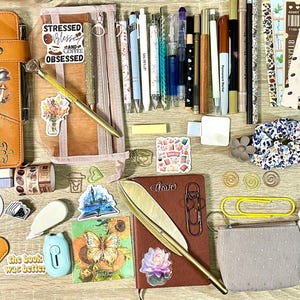 May include: A collection of stationery items including a tan leather journal with a tree design, a brown notebook, various pens, markers, bookmarks, stickers, a floral scrunchie, a small pouch, paper clips, and reading list inserts arranged on a flat surface.