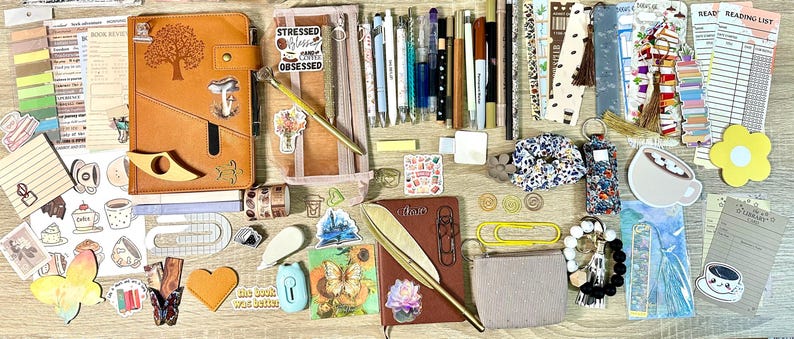 May include: A collection of stationery items including a tan leather journal with a tree design, a brown notebook, various pens, markers, bookmarks, stickers, a floral scrunchie, a small pouch, paper clips, and reading list inserts arranged on a flat surface.