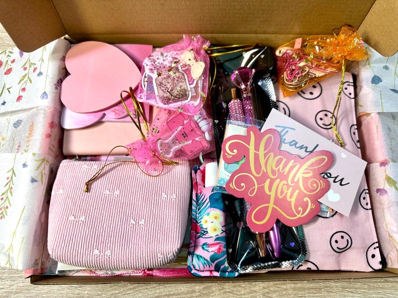 Pu&ograve; includere: Una scatola regalo in cartone riempita di articoli di cancelleria e accessori, tra cui una pochette in velluto a coste rosa, foglietti adesivi a forma di cuore, un astuccio a fiori, penne con tappi a diamante, un biglietto di ringraziamento e sacchetti.
