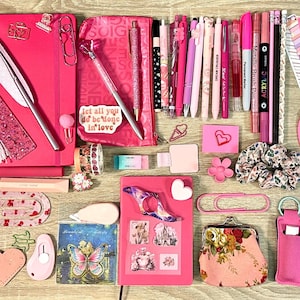 Pu&ograve; includere: Una collezione di articoli di cancelleria rosa che include quaderni, penne, pennarelli, graffette, adesivi, segnalibri, un piccolo portamonete, un elastico per capelli floreale e vari foglietti adesivi disposti su una superficie.