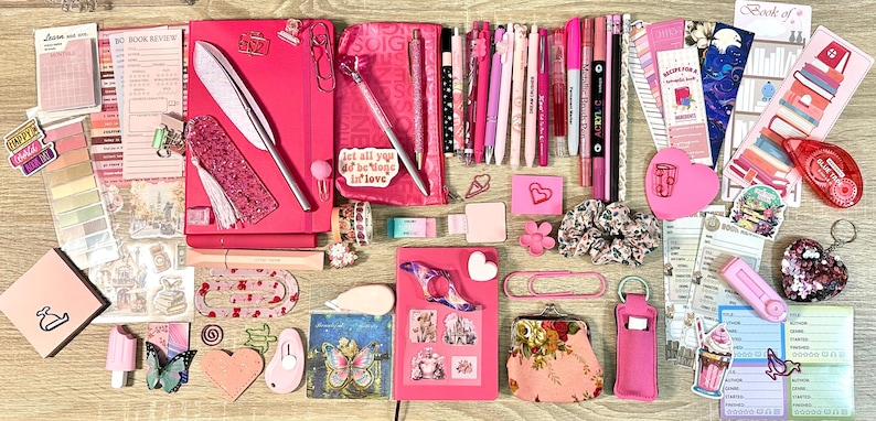 Pu&ograve; includere: Una collezione di articoli di cancelleria rosa che include quaderni, penne, pennarelli, graffette, adesivi, segnalibri, un piccolo portamonete, un elastico per capelli floreale e vari foglietti adesivi disposti su una superficie.