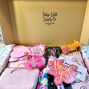 Pu&ograve; includere: Una scatola di cartone con l'etichetta Indigo Wild Supply Co contenente vari articoli, tra cui un blocco note rosa a forma di cuore, una custodia a righe rosa, una custodia con motivo floreale, un biglietto di ringraziamento e sacchetti regalo.