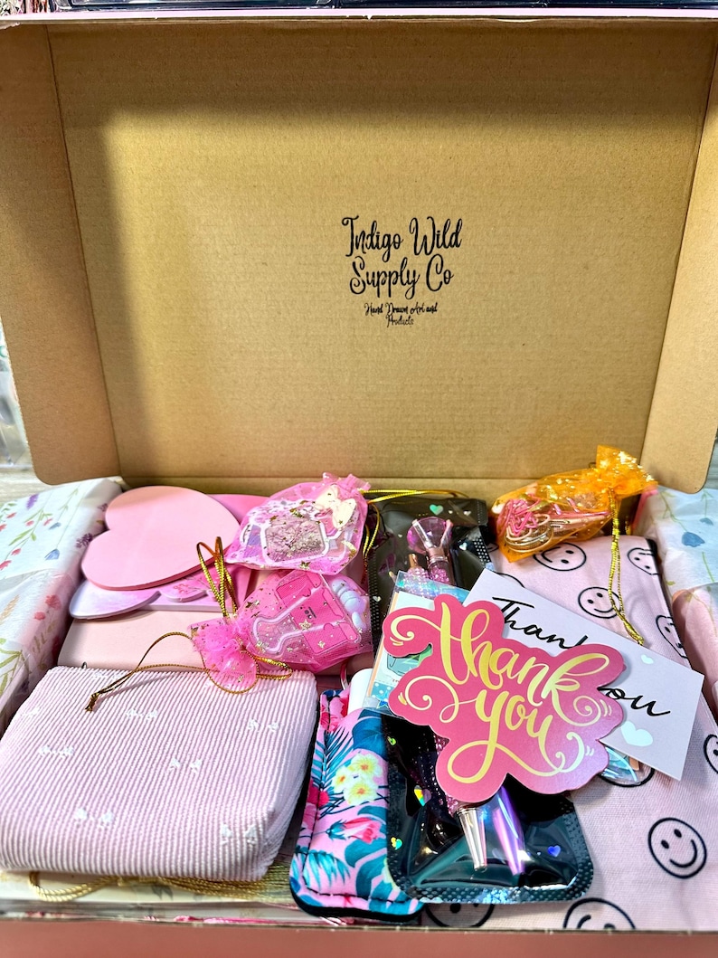 Pu&ograve; includere: Una scatola di cartone con l'etichetta Indigo Wild Supply Co contenente vari articoli, tra cui un blocco note rosa a forma di cuore, una custodia a righe rosa, una custodia con motivo floreale, un biglietto di ringraziamento e sacchetti regalo.