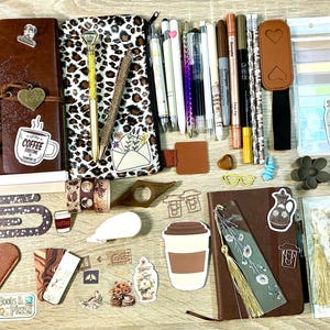 May include: A collection of stationery items including two leather-bound journals, a leopard print pencil case, various pens, markers, and highlighters. Also included are decorative stickers, bookmarks, a gold key, a scrunchie, and paper clips.