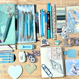 Può includere: Una collezione di articoli di cancelleria nelle tonalità del blu e del verde acqua, tra cui un diario, penne, segnalibri, adesivi, nastro washi, un astuccio, graffette, un portachiavi e un elastico per capelli, disposti su una superficie in legno chiaro.