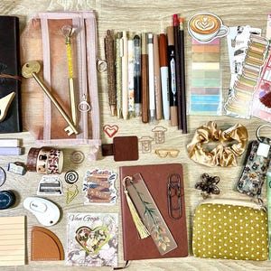 May include: A collection of stationery items including a dark leather journal, a brown notebook, various pens, pencils, markers, a mesh pencil case, a gold key-shaped pen, a gold scrunchie, a polka dot pouch, bookmarks, stickers, and reading lists.