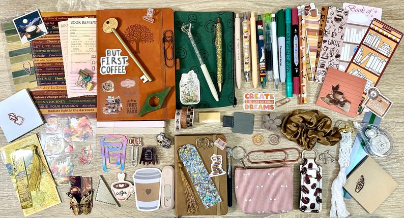 May include: A collection of stationery items including two notebooks, various pens, markers, bookmarks, stickers, a small pouch, a scrunchie, and paper clips. The items feature coffee-themed designs, floral patterns, and motivational text.