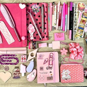 Pu&ograve; includere: Una collezione di articoli di cancelleria rosa e viola che include due quaderni, un astuccio, varie penne, pennarelli, foglietti adesivi, graffette, un elastico per capelli, un piccolo portamonete e adesivi decorativi con libri, fiori e farfalle.