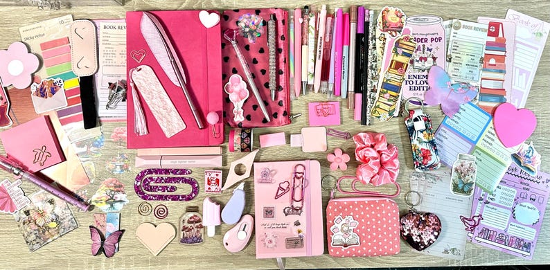 Pu&ograve; includere: Una collezione di articoli di cancelleria rosa e viola che include due quaderni, un astuccio, varie penne, pennarelli, foglietti adesivi, graffette, un elastico per capelli, un piccolo portamonete e adesivi decorativi con libri, fiori e farfalle.
