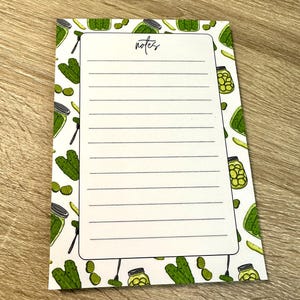 Può includere: Un blocco note rettangolare con un centro bianco con linee orizzontali e la parola notes stampata in alto. Il bordo è decorato con un motivo ripetitivo di cetriolini verdi, fette di cetriolino e barattoli di vetro pieni di cetriolini.