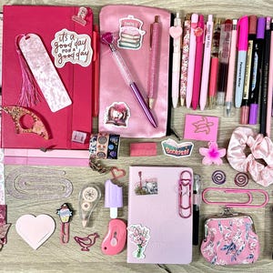 Pu&ograve; includere: Una collezione di articoli di cancelleria rosa e magenta che comprende due quaderni, un astuccio, varie penne, pennarelli, foglietti adesivi, graffette, un elastico per capelli, un piccolo portamonete e adesivi decorativi con motivi floreali e letterari.