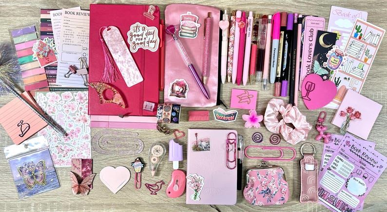 Pu&ograve; includere: Una collezione di articoli di cancelleria rosa e magenta che comprende due quaderni, un astuccio, varie penne, pennarelli, foglietti adesivi, graffette, un elastico per capelli, un piccolo portamonete e adesivi decorativi con motivi floreali e letterari.
