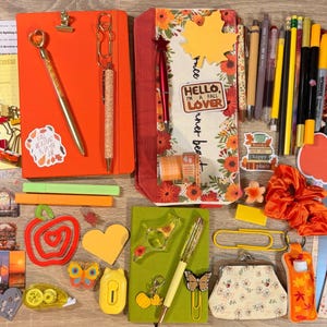 May include: A collection of stationery items including an orange notebook, a floral pencil case, various pens, markers, paper clips, sticky notes, and stickers with autumn themes, including one sticker that reads HELLO I AM A FALL LOVER.