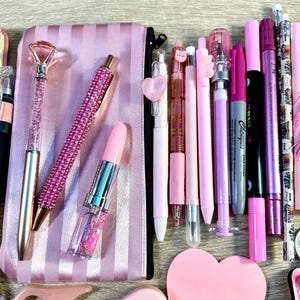 Pu&ograve; includere: Una collezione di articoli di cancelleria rosa che include un astuccio a righe, una penna con punta a diamante, una penna con strass, una penna a forma di rossetto, varie penne gel, pennarelli, matite e un blocco note a forma di cuore.