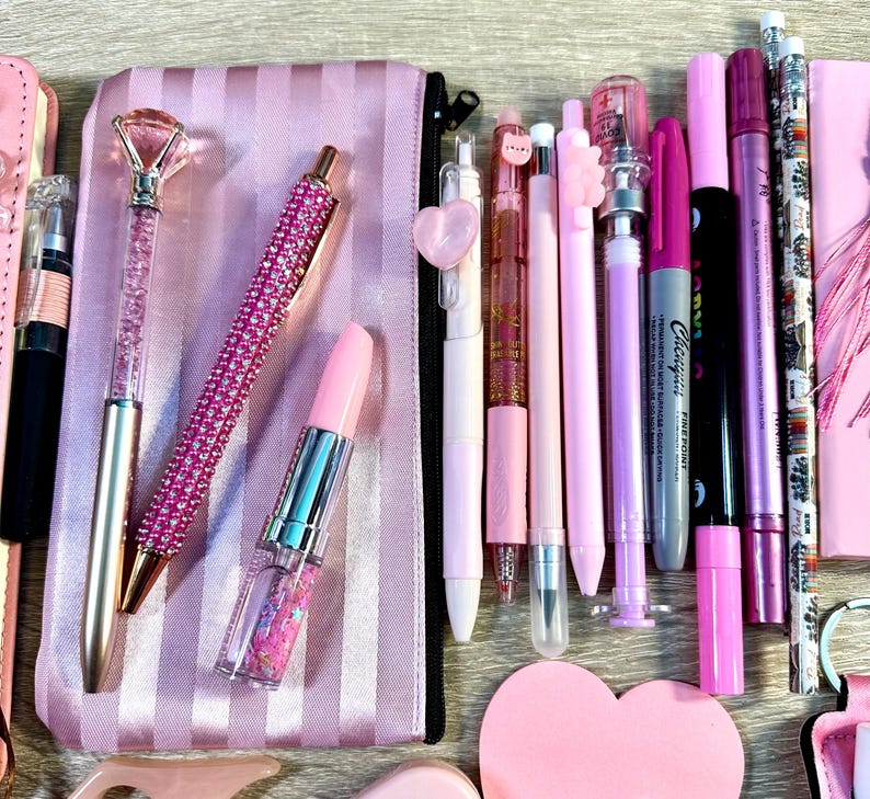 Pu&ograve; includere: Una collezione di articoli di cancelleria rosa che include un astuccio a righe, una penna con punta a diamante, una penna con strass, una penna a forma di rossetto, varie penne gel, pennarelli, matite e un blocco note a forma di cuore.