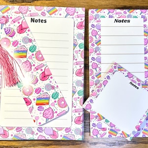 May include: A stationery set featuring a notebook, notepad, and bookmark. The items are decorated with a colorful candy pattern, including cupcakes, donuts, and cotton candy. The bookmark has a pink tassel. The notebook and notepad have the word "Notes" printed on them.