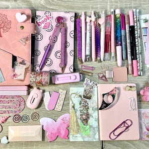Pu&ograve; includere: Una collezione di articoli di cancelleria rosa che comprende due quaderni, varie penne, foglietti adesivi, segnalibri, graffette, una piccola custodia, un biglietto a forma di farfalla, un elastico per capelli e adesivi decorativi.