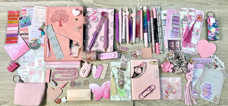 Pu&ograve; includere: Una collezione di articoli di cancelleria rosa che comprende due quaderni, varie penne, foglietti adesivi, segnalibri, graffette, una piccola custodia, un biglietto a forma di farfalla, un elastico per capelli e adesivi decorativi.
